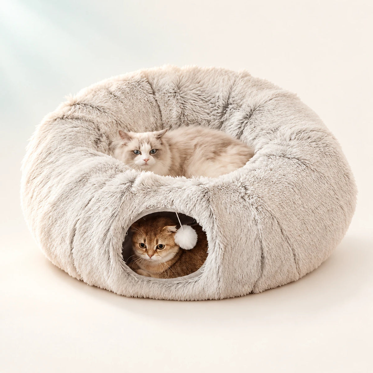 Two cats in a cream round tunnel bed on a warm ivory background.